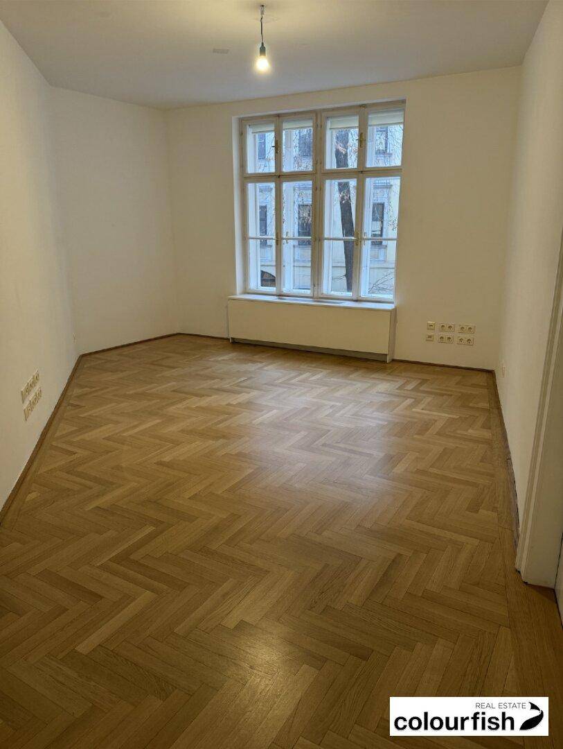 Heller Raum mit klassischem Fischgrätparkett, weißen Wänden und einem großen Fenster, das den Raum erhellt.