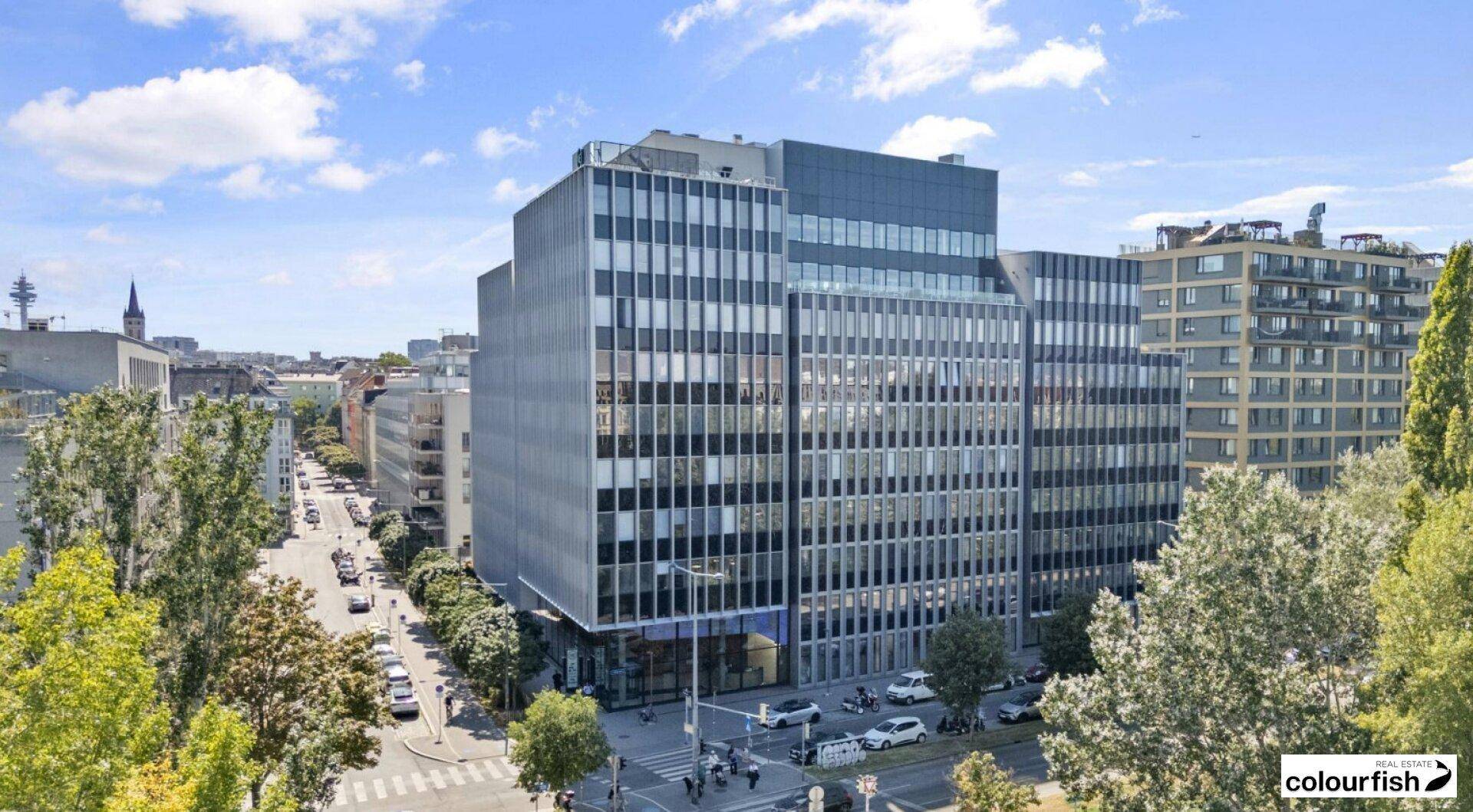 Modernes Bürogebäude mit markanter Glasfassade in einer belebten städtischen Umgebung, umgeben von Bäumen und anderen Gebäuden.