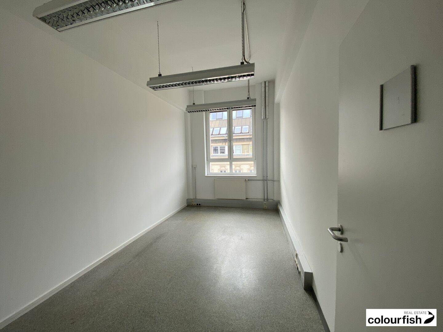 Leerer Büroraum mit einem Fenster, das Tageslicht hereinlässt und einfachem Bodenbelag.