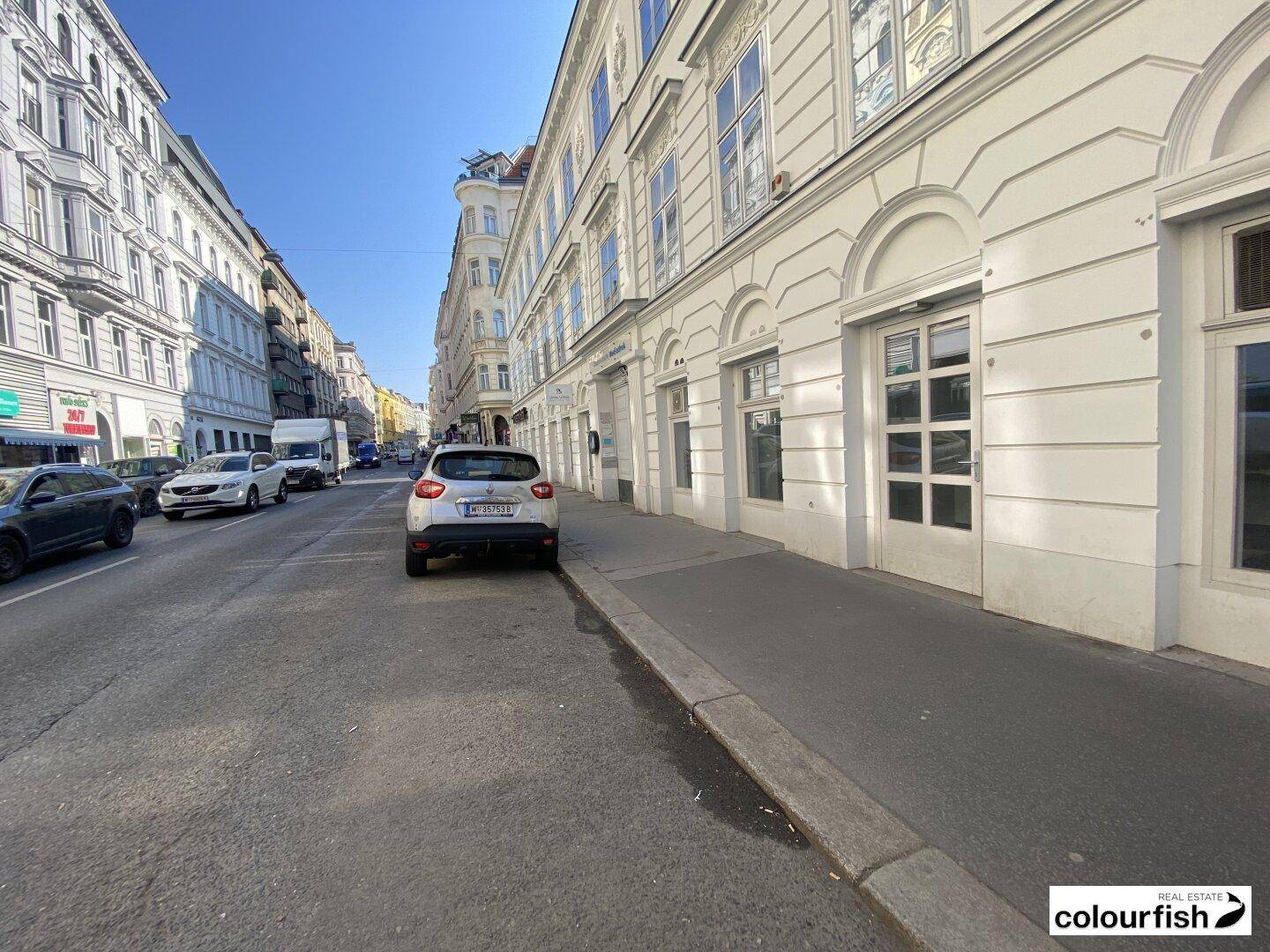 Blick die Straße entlang mit parkenden Autos und historischen Gebäuden unter blauem Himmel.