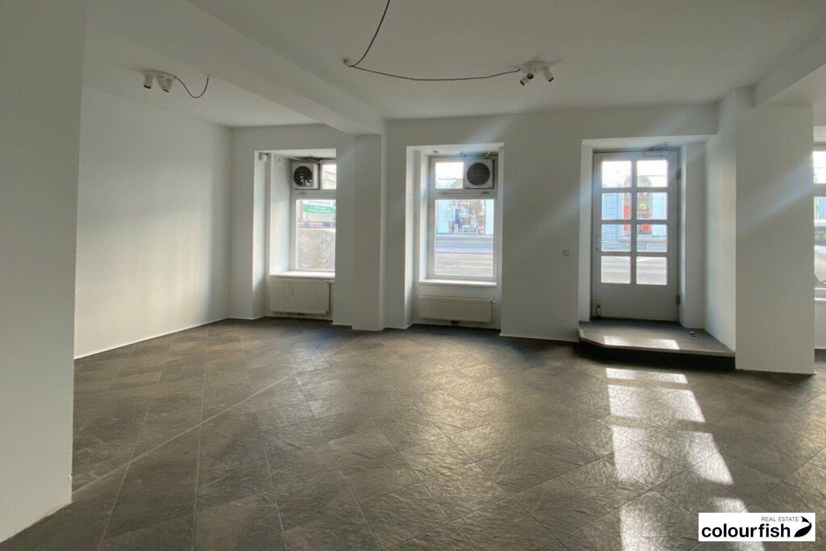 Leerer Gewerberaum mit dunklen Fliesen, weißen Wänden und großen Fenstern zur Straße.