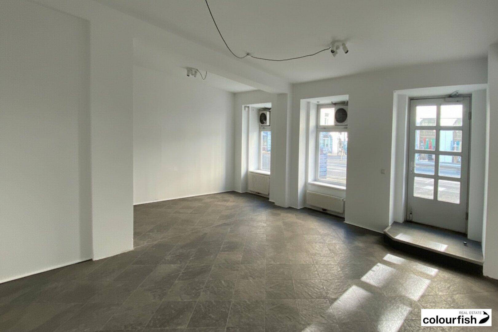 Geräumiger Gewerberaum mit Fliesenboden, hellen Wänden und viel Tageslicht durch große Fenster.