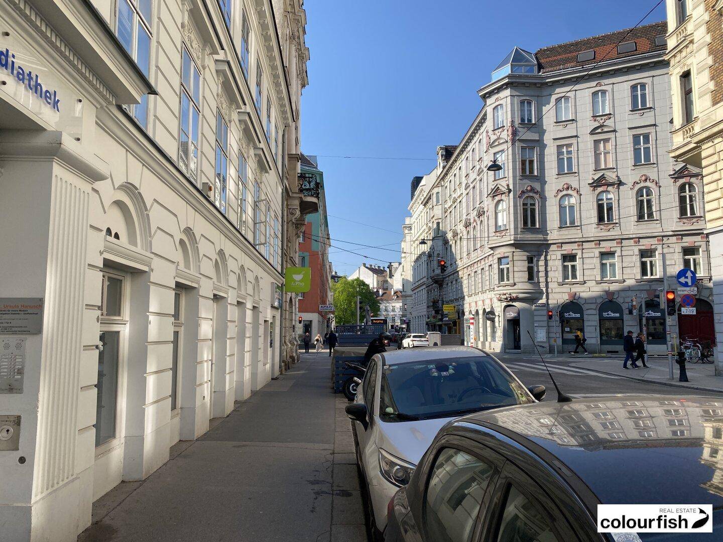 Städtische Straßenszene mit Gebäuden, parkenden Autos und Fußgängern an einem sonnigen Tag.