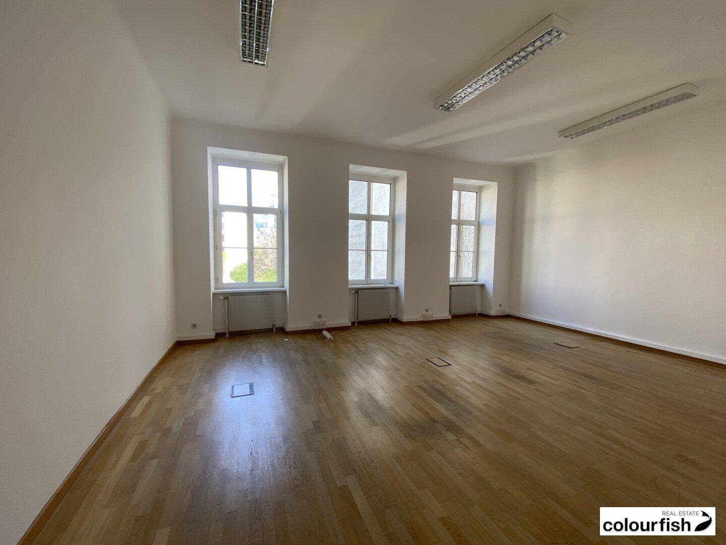 Geräumiges Büro mit hellem Parkettboden und großen Fenstern, die viel Tageslicht hereinlassen.
