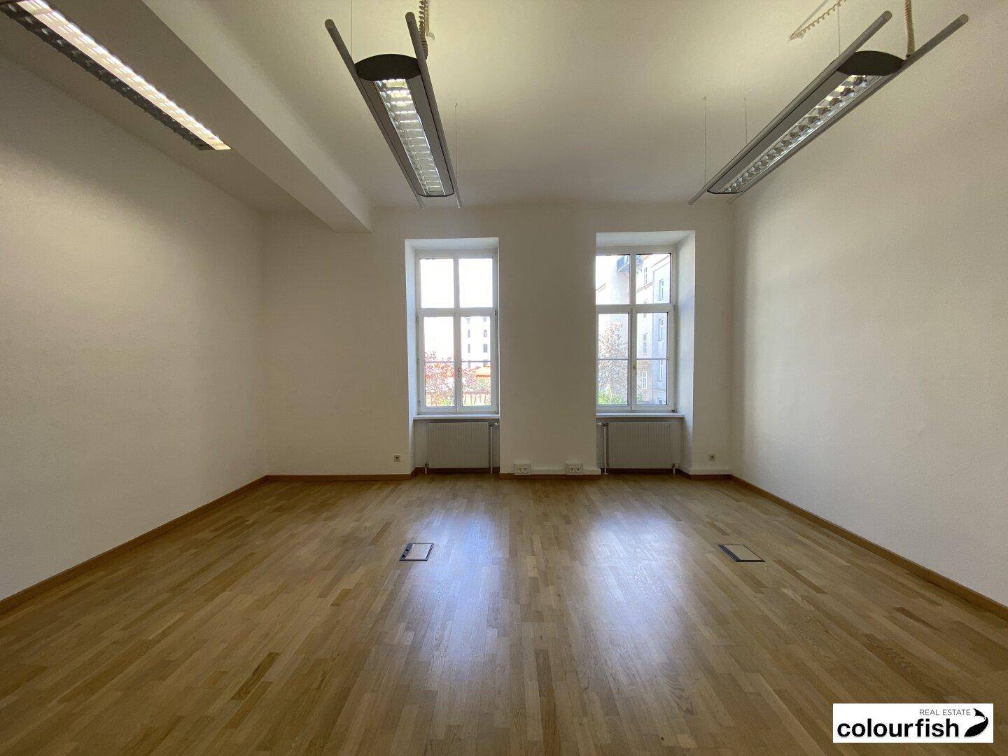 Modernes Büro mit Holzboden und zwei großen Fenstern, die den Raum mit Licht füllen.