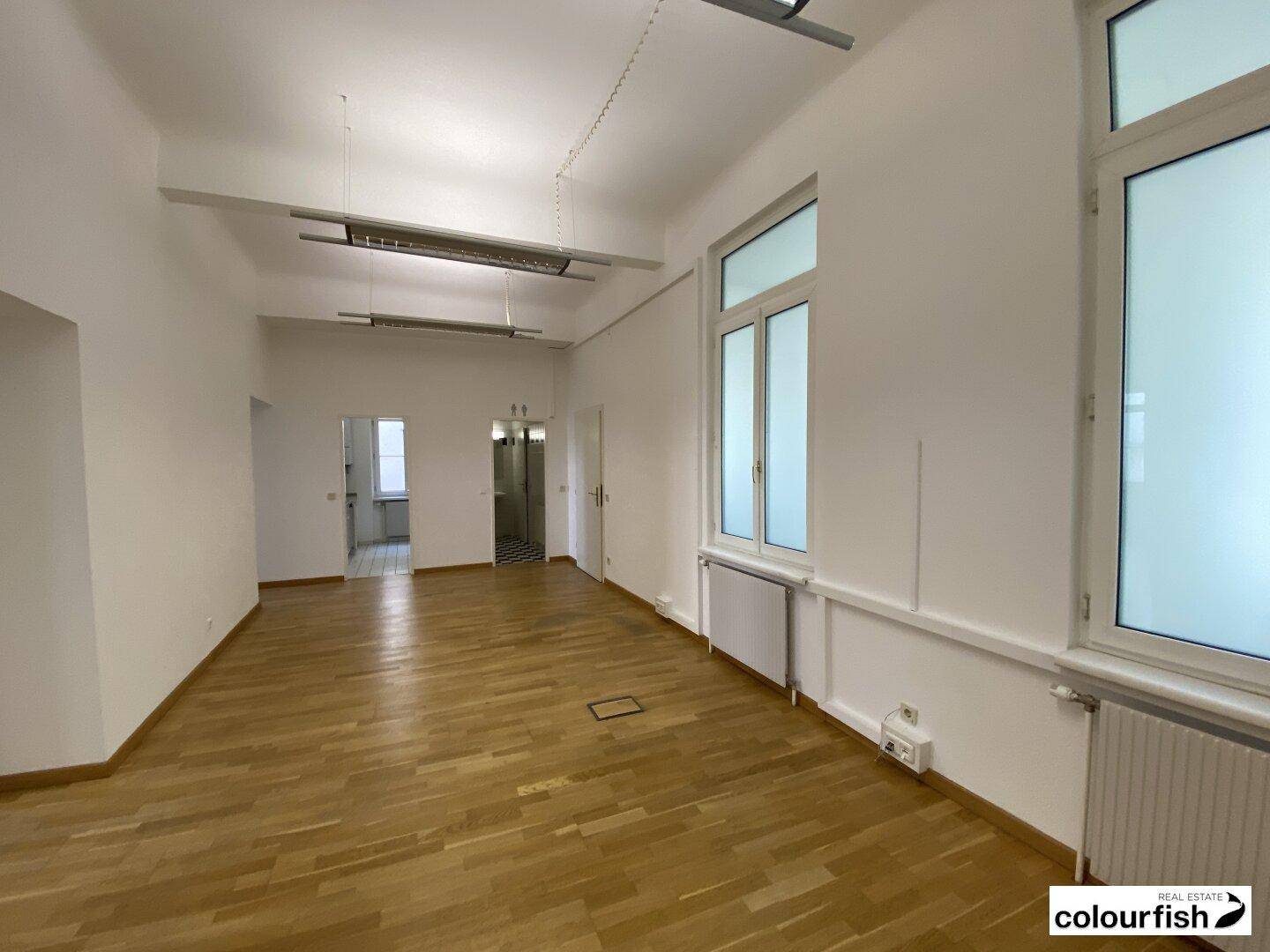 Großzügiger Büroraum mit Holzboden und hohen Fenstern, der einen Blick in angrenzende Bereiche bietet.