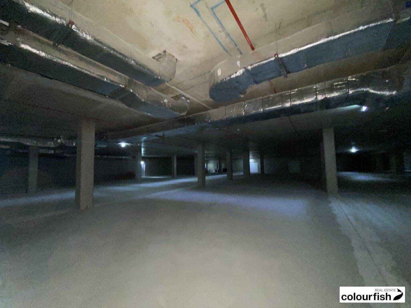 Weitläufige Tiefgarage mit Betonboden, tragenden Säulen und freiliegenden Lüftungsleitungen unter der Decke.