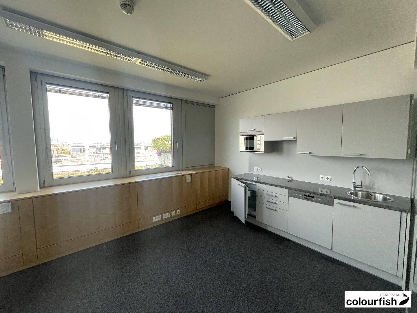 Moderne Küchenzeile in einem Büroraum mit grauen Schränken, Spüle und integrierten Geräten.