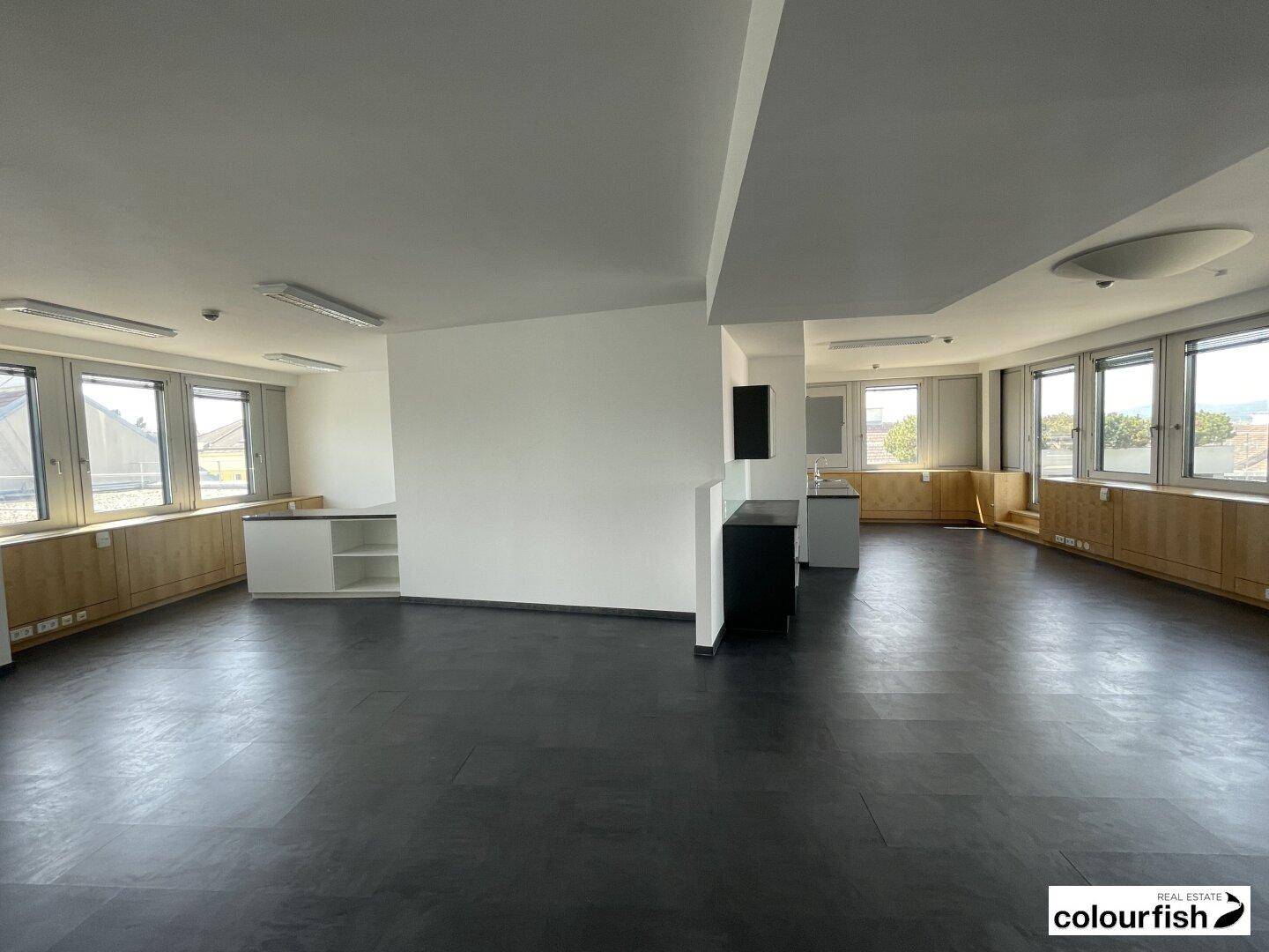 Offener Bürobereich mit dunklem Vinylboden, weißen Wänden und mehreren Fenstern.
