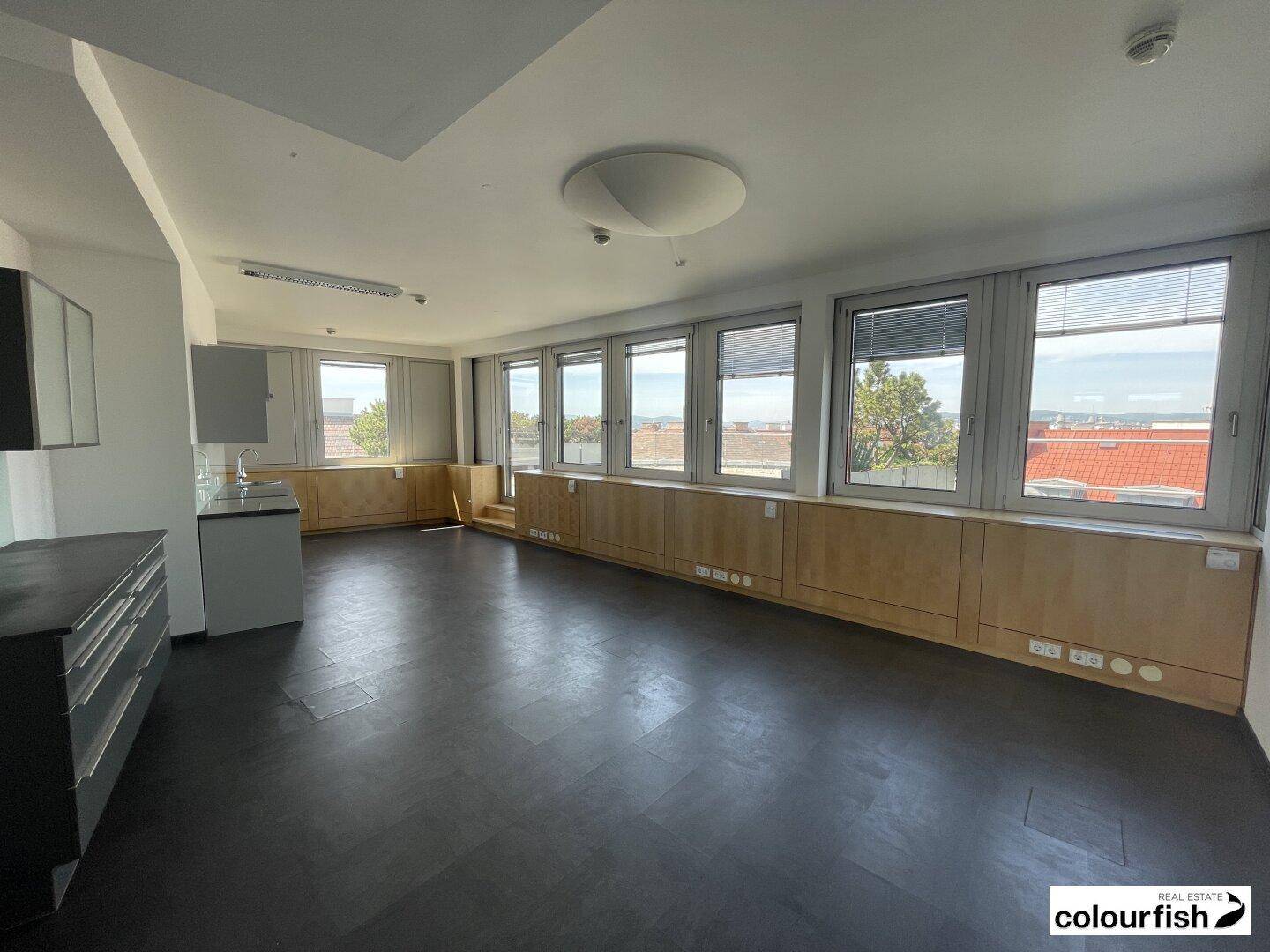 Heller Büroraum mit dunklem Vinylboden, vielen Fenstern und einer kleinen Küchenzeile.