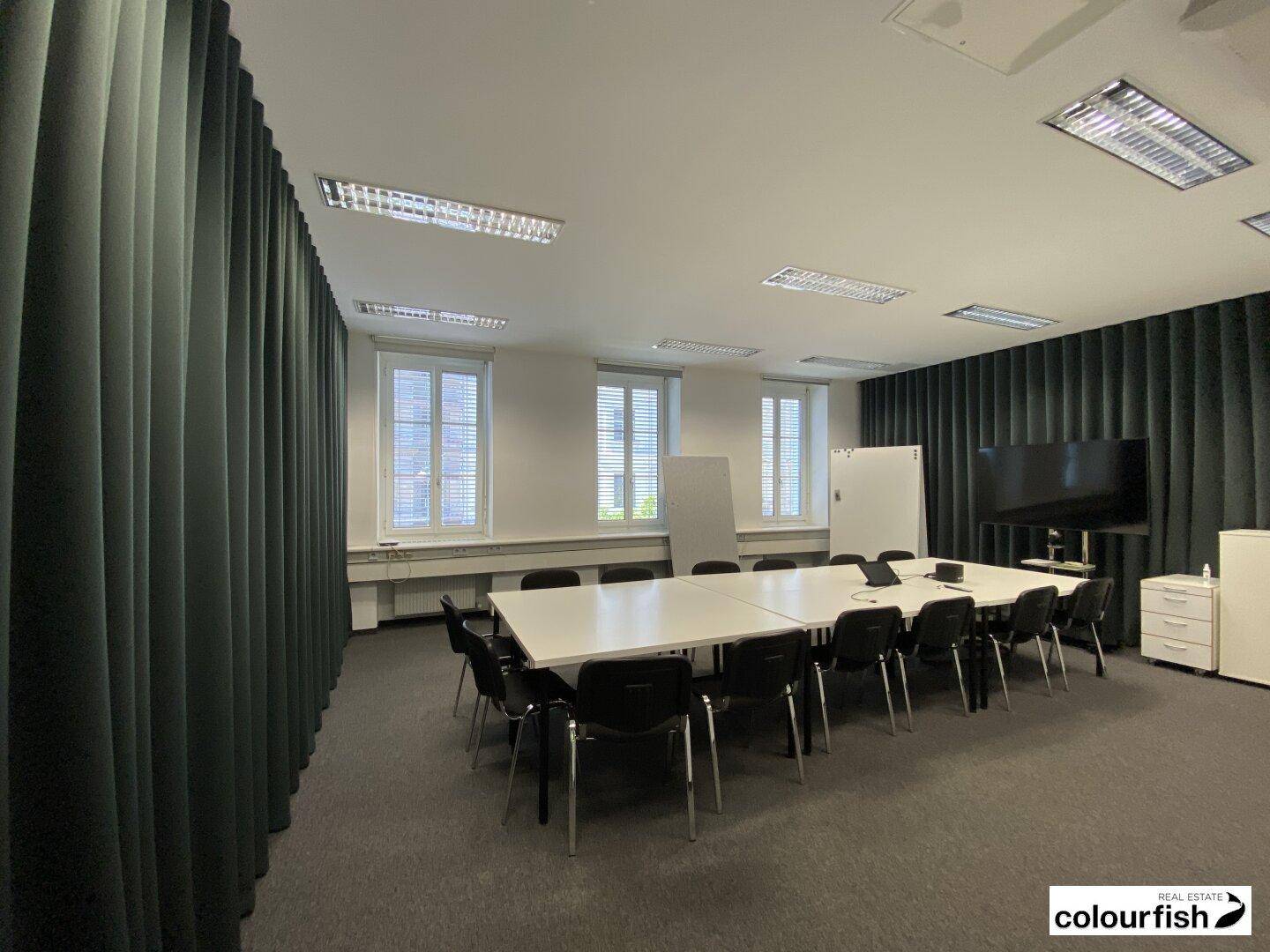 Großer Konferenzraum mit langem Tisch, vielen Stühlen und einem Bildschirm für Präsentationen.