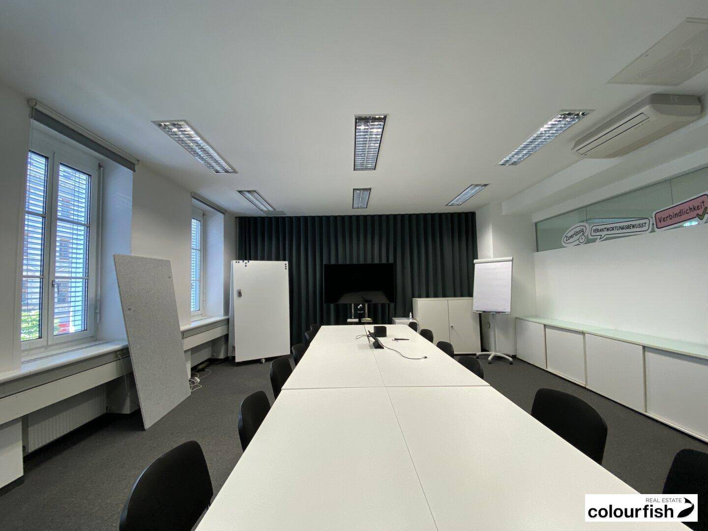 Geräumiger Konferenzraum mit großem Tisch, Stühlen, Whiteboards und einem Bildschirm für Besprechungen.