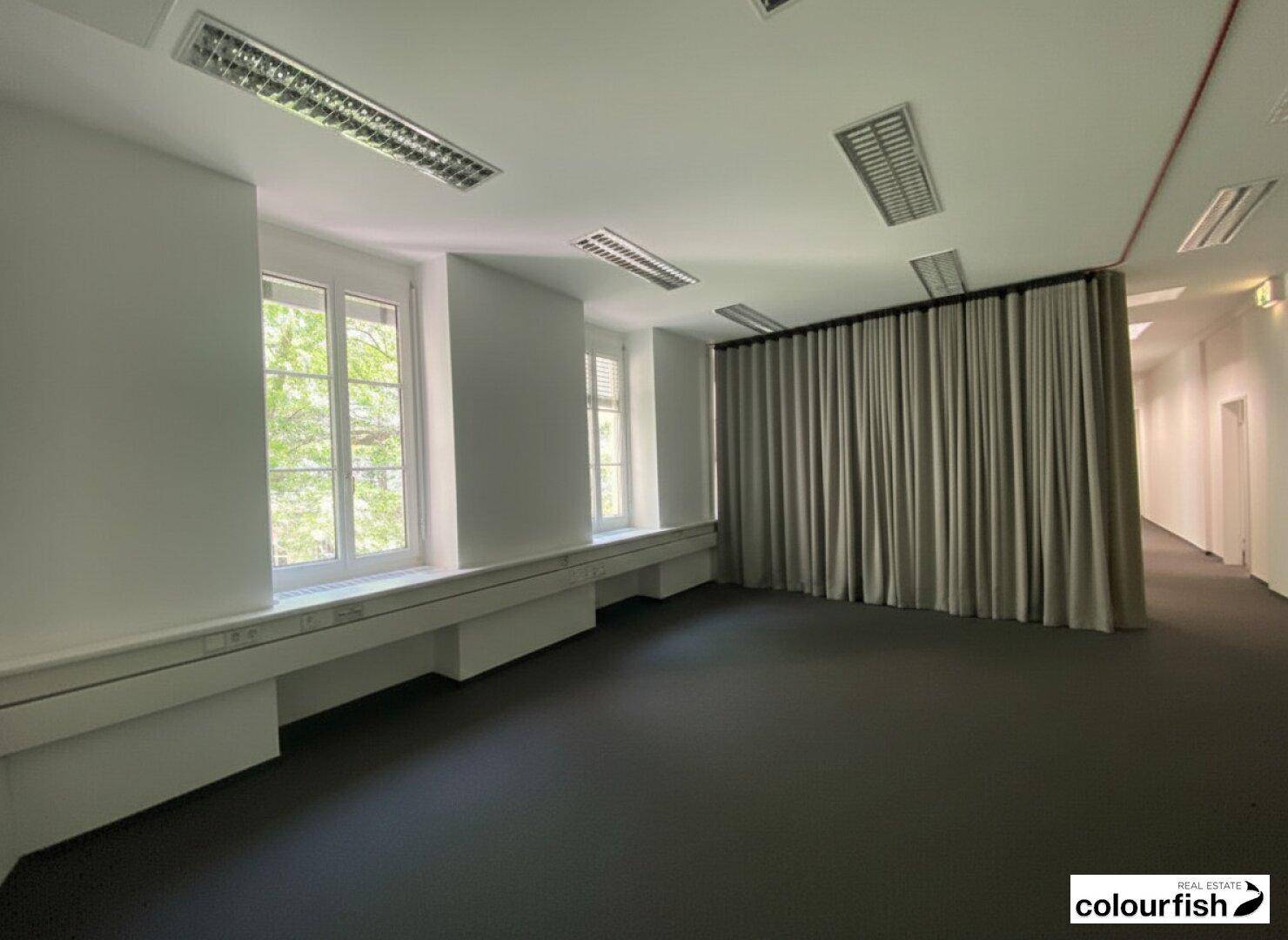 Helles Büro mit Teppichboden, großen Fenstern und einem Vorhang, der den Raum abtrennt, sowie einem Flur im Hintergrund.