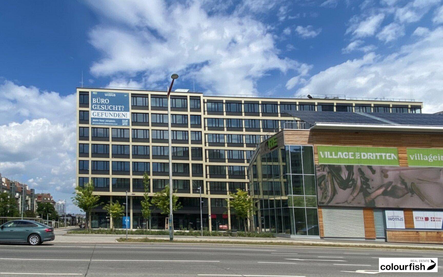 Modernes Bürogebäude mit vielen Fenstern und einer belebten Straße im Vordergrund unter blauem Himmel.