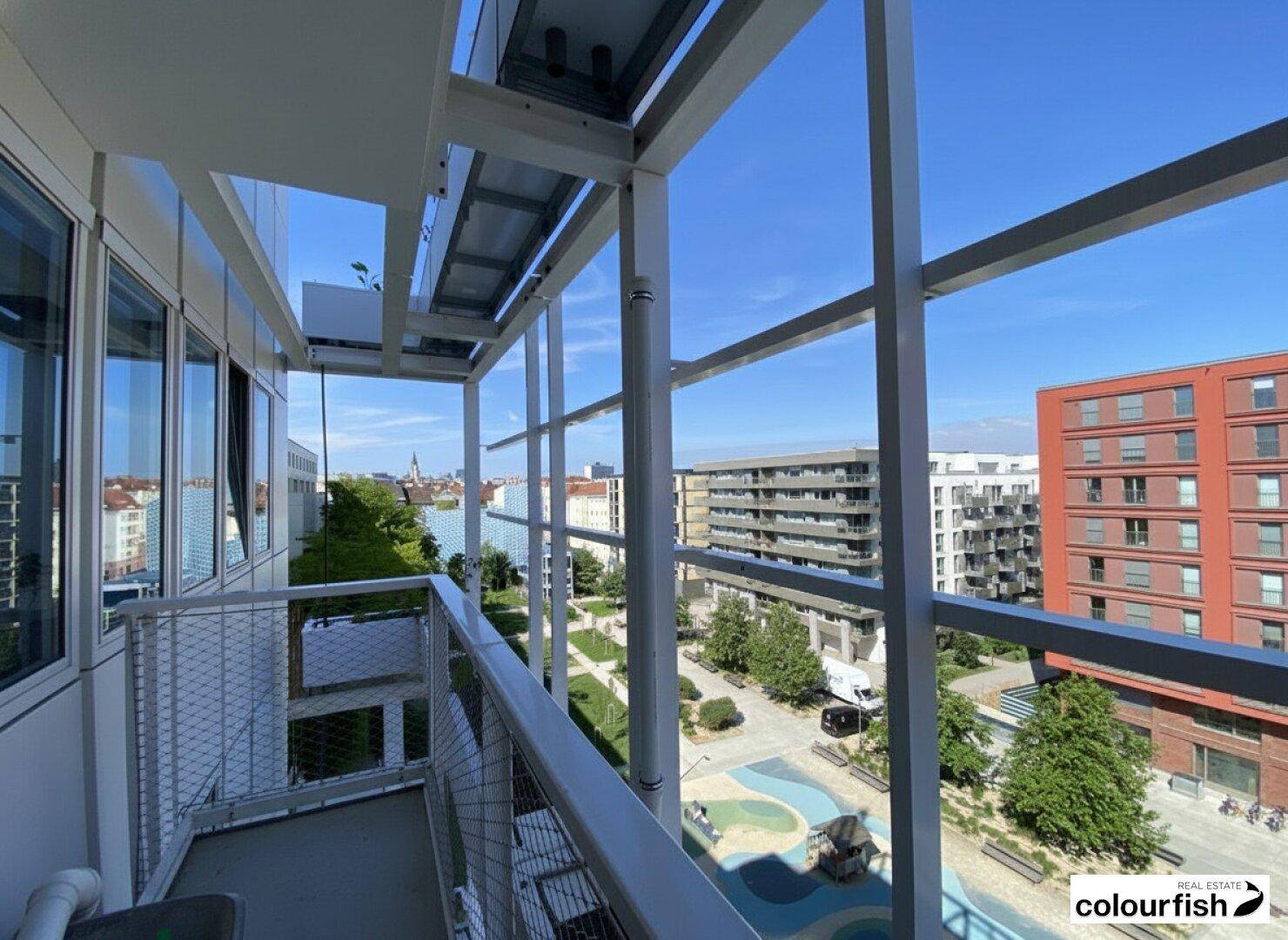 Blick vom modernen Balkon auf den grünen Innenhof und die umliegenden Gebäude unter blauem Himmel.