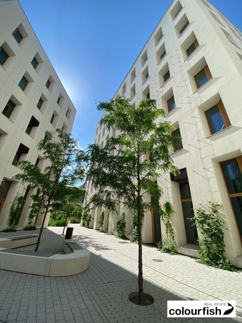 Moderner Innenhof zwischen weißen Gebäuden mit Bäumen und gepflasterten Wegen unter blauem Himmel.