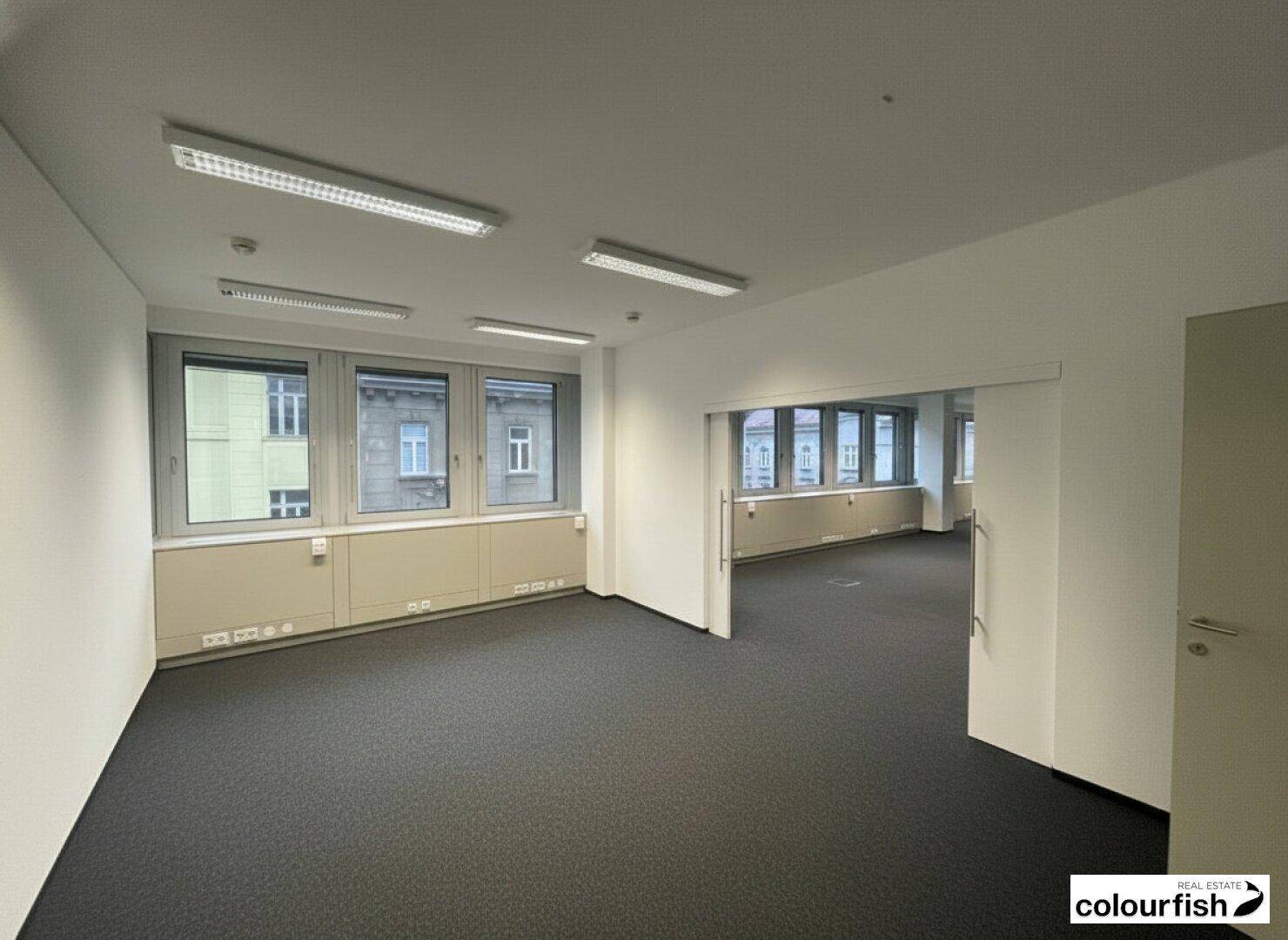 Heller Bürobereich mit Teppichboden, großen Fenstern und Zugang zu weiteren Räumen.