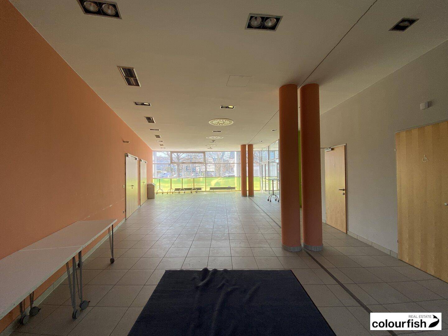 Großer, heller Saal mit Fliesenboden, hohen Decken und großen Fenstern, ideal für kommerzielle Nutzung oder Veranstaltungen.