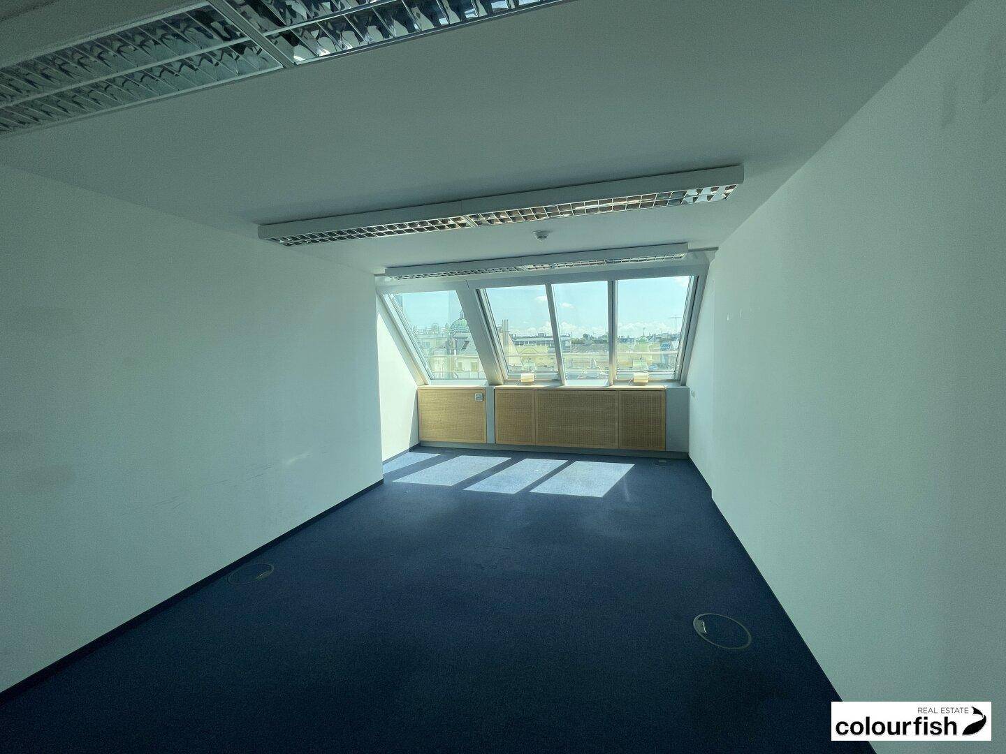 Büro mit schrägen Fenstern, blauem Teppichboden und Blick auf die Umgebung, gut beleuchtet.