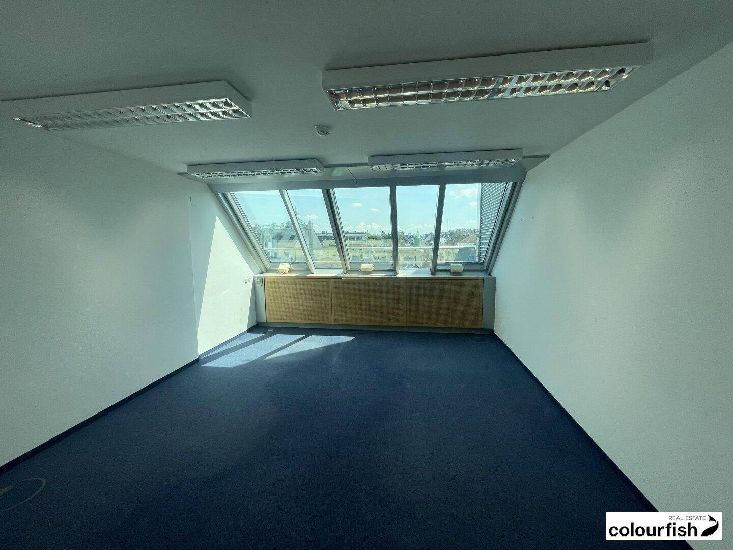Geräumiges Büro mit schrägen Fenstern, blauem Teppichboden und viel Tageslicht, ideal für produktives Arbeiten.