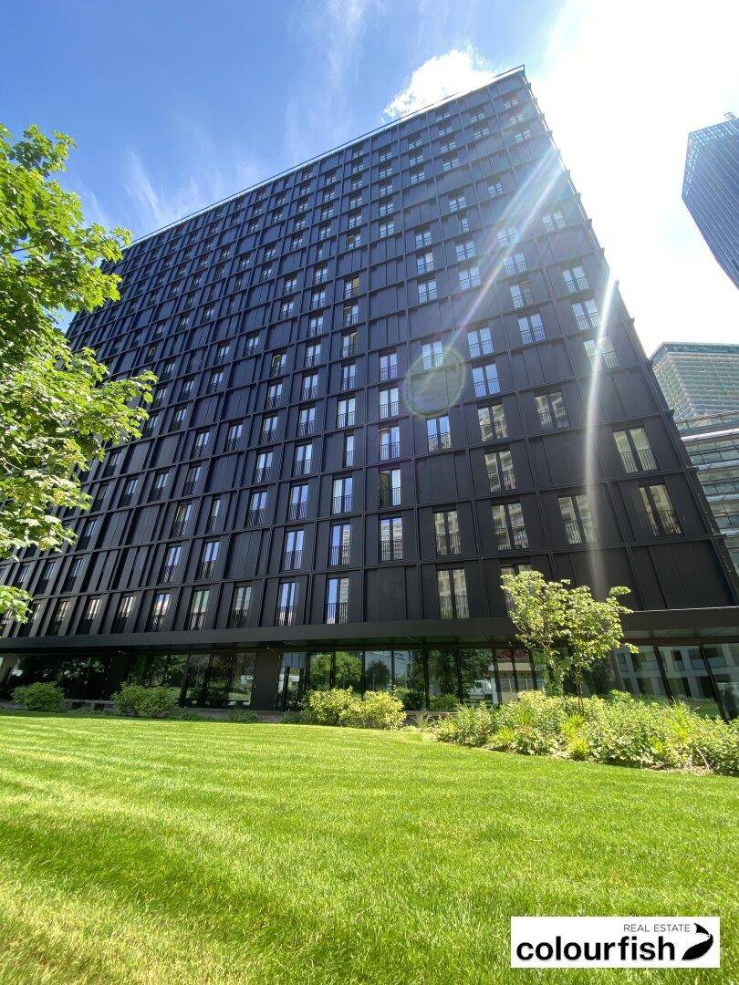 Sonnenbeschienene moderne Gebäudefassade mit dunklen Elementen und großen Fenstern, umgeben von Grün.