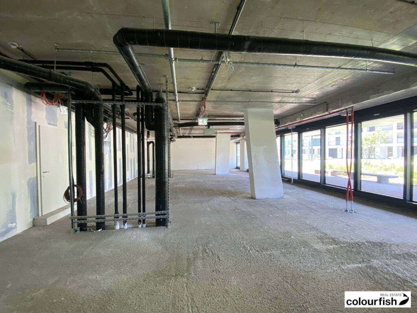 Weiterer Blick in den unfertigen Gewerberaum mit vielen sichtbaren Rohren, Betonflächen und großen Fenstern.