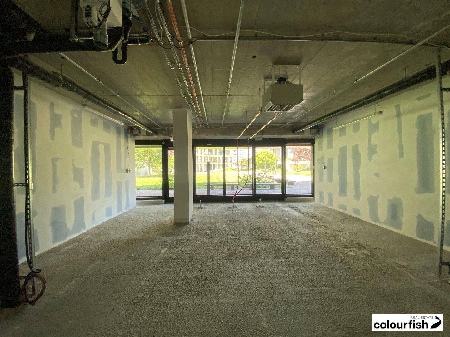 Rohbau eines Innenraums mit großen Fenstern, Betonboden und sichtbaren Installationen, viel Tageslicht.