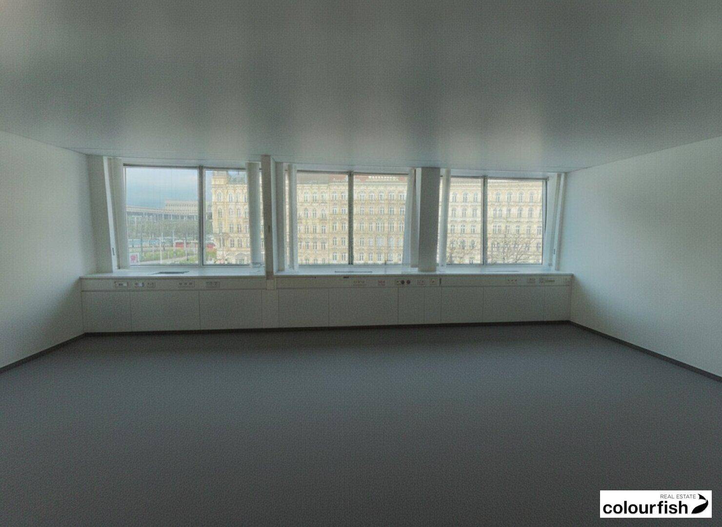 Leerer Raum mit drei großen Fenstern, die einen Blick auf die Stadtgebäude bieten und viel Tageslicht hereinlassen.