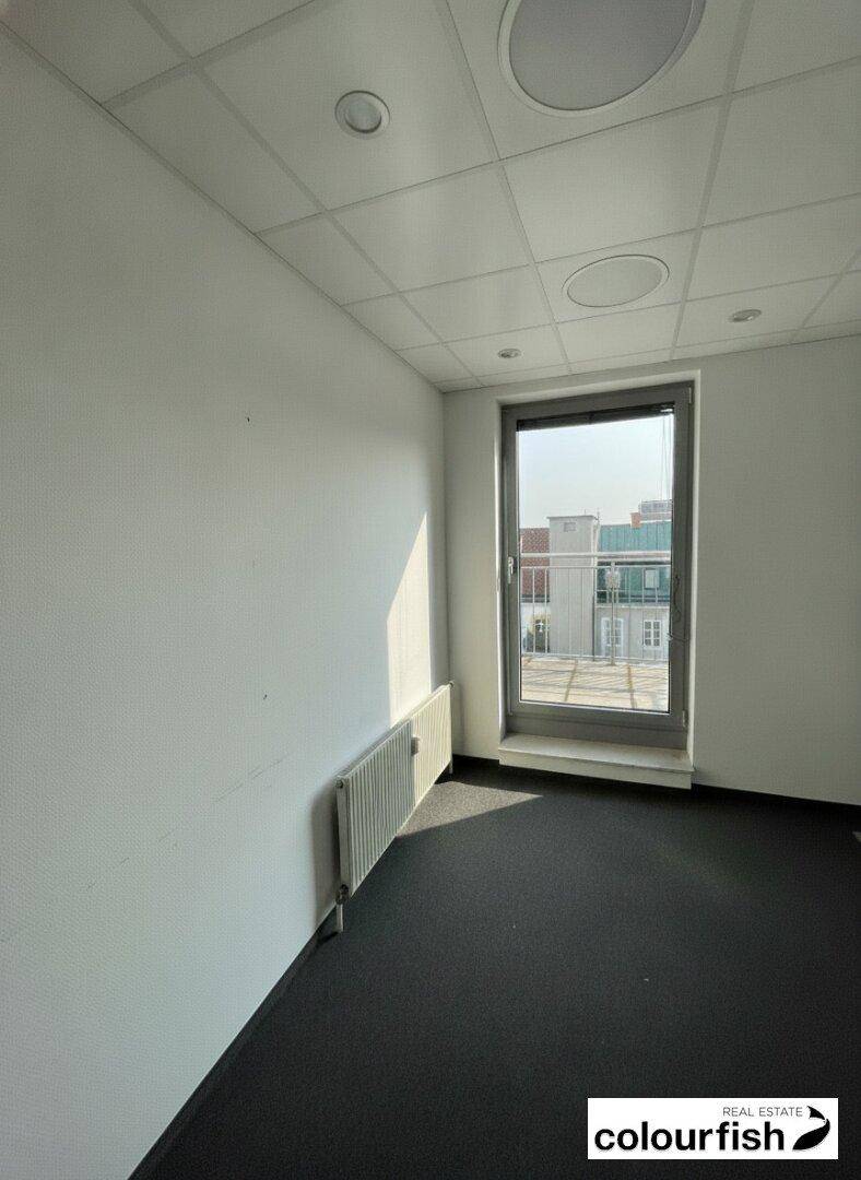 Heller Büroraum mit Teppichboden und Blick auf umliegende Gebäude.