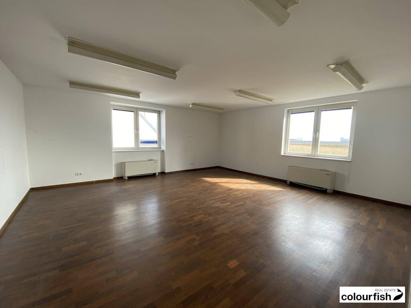 Heller, leerer Büroraum mit dunklem Holzfußboden, weißen Wänden und zwei großen Fenstern.