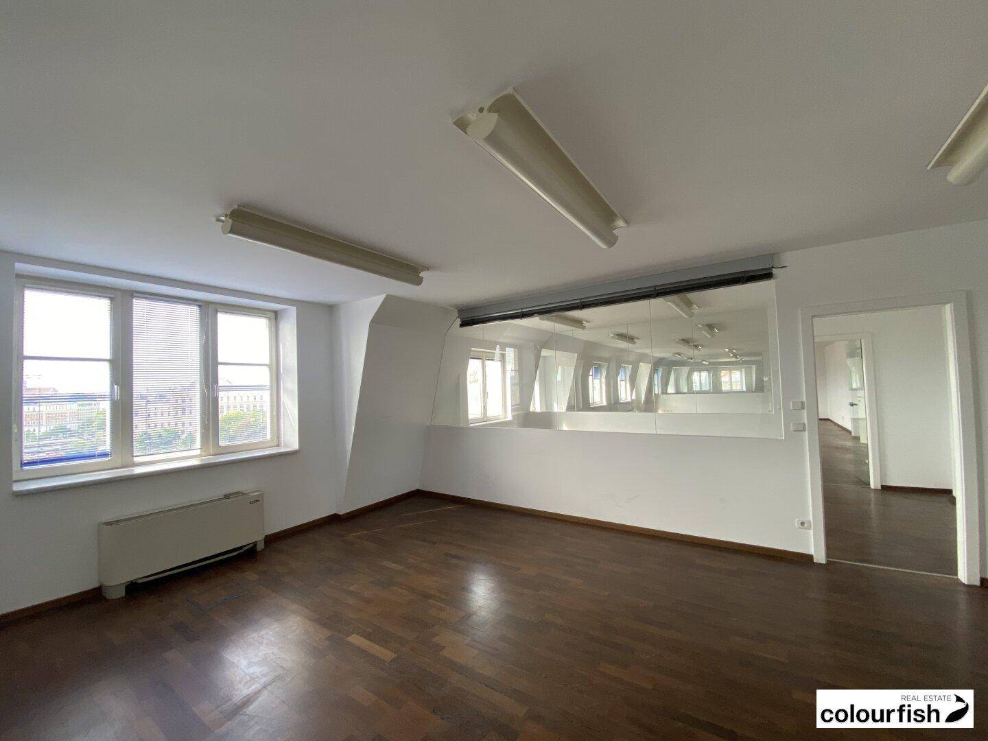 Heller Büroraum mit dunklem Holzfußboden, weißen Wänden und einem Fenster mit Jalousien.