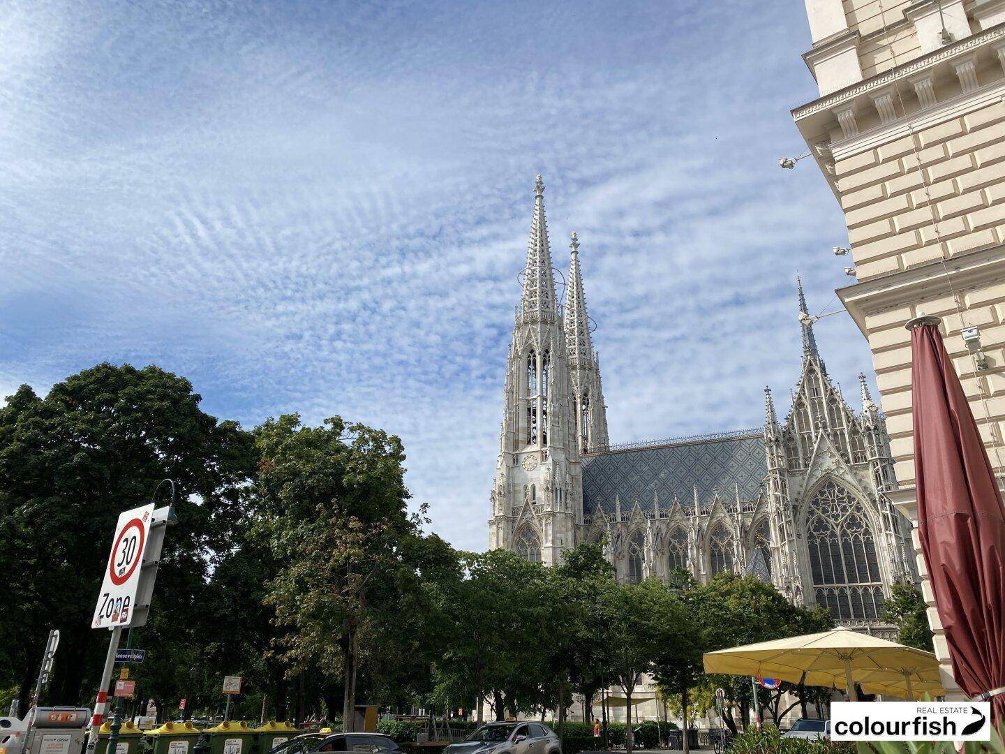 Blick auf eine imposante gotische Kirche von der Straße aus, umgeben von Bäumen und Stadtleben.