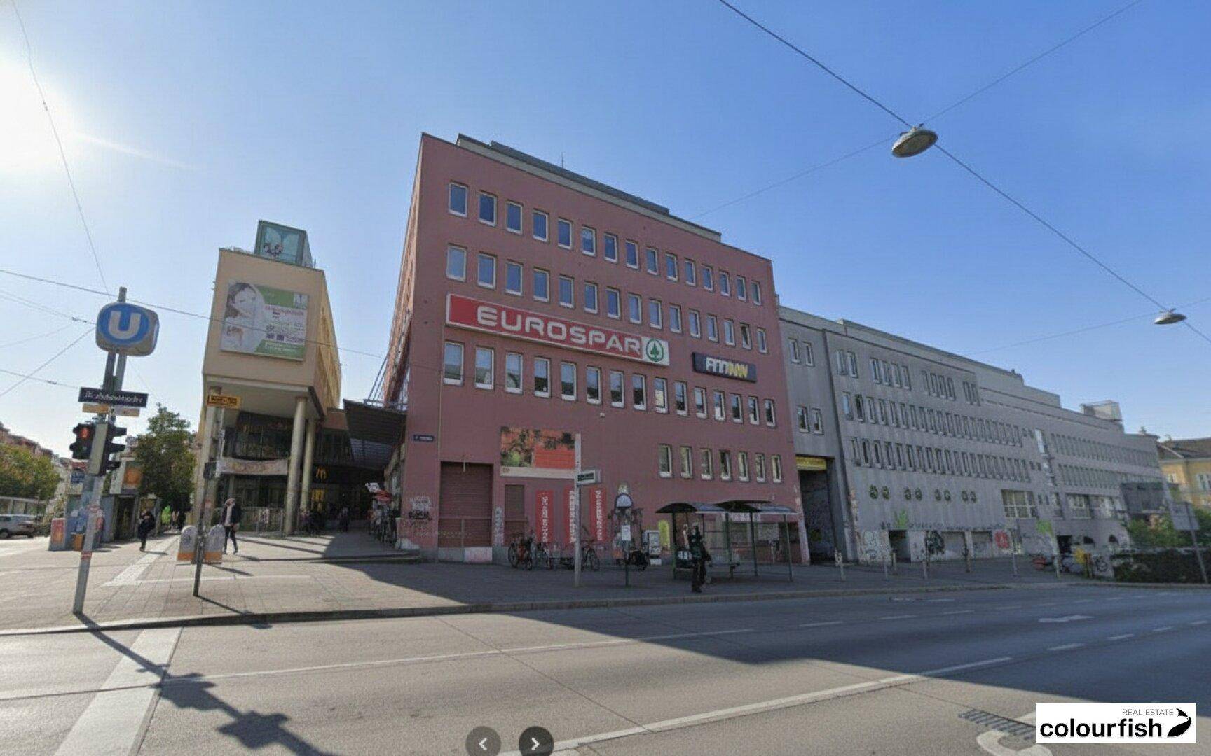 Ansicht eines Geschäftsgebäudes mit Supermarkt und Fitnessstudio an einer belebten Straße.