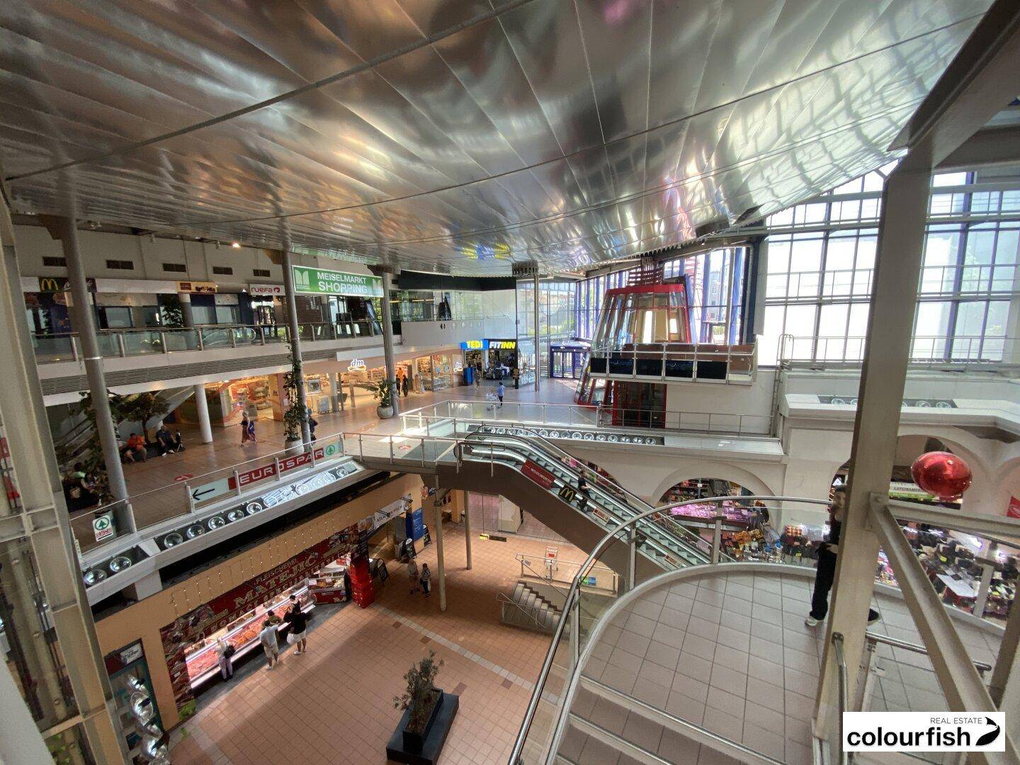 Großes, mehrstöckiges Einkaufszentrum mit Rolltreppen, Geschäften und viel Tageslicht durch Glasfassade.