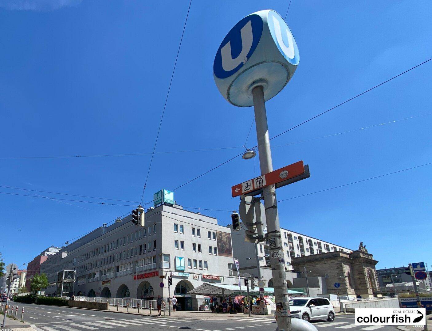 Außenansicht einer belebten Straße mit U-Bahn-Station, Gebäuden und klarem blauen Himmel.
