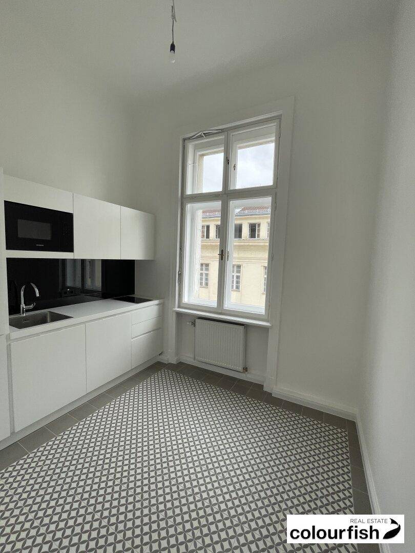 Kleine Küchenzeile mit weißen Schränken, schwarzer Rückwand, integrierter Mikrowelle und gemusterten Fliesen.