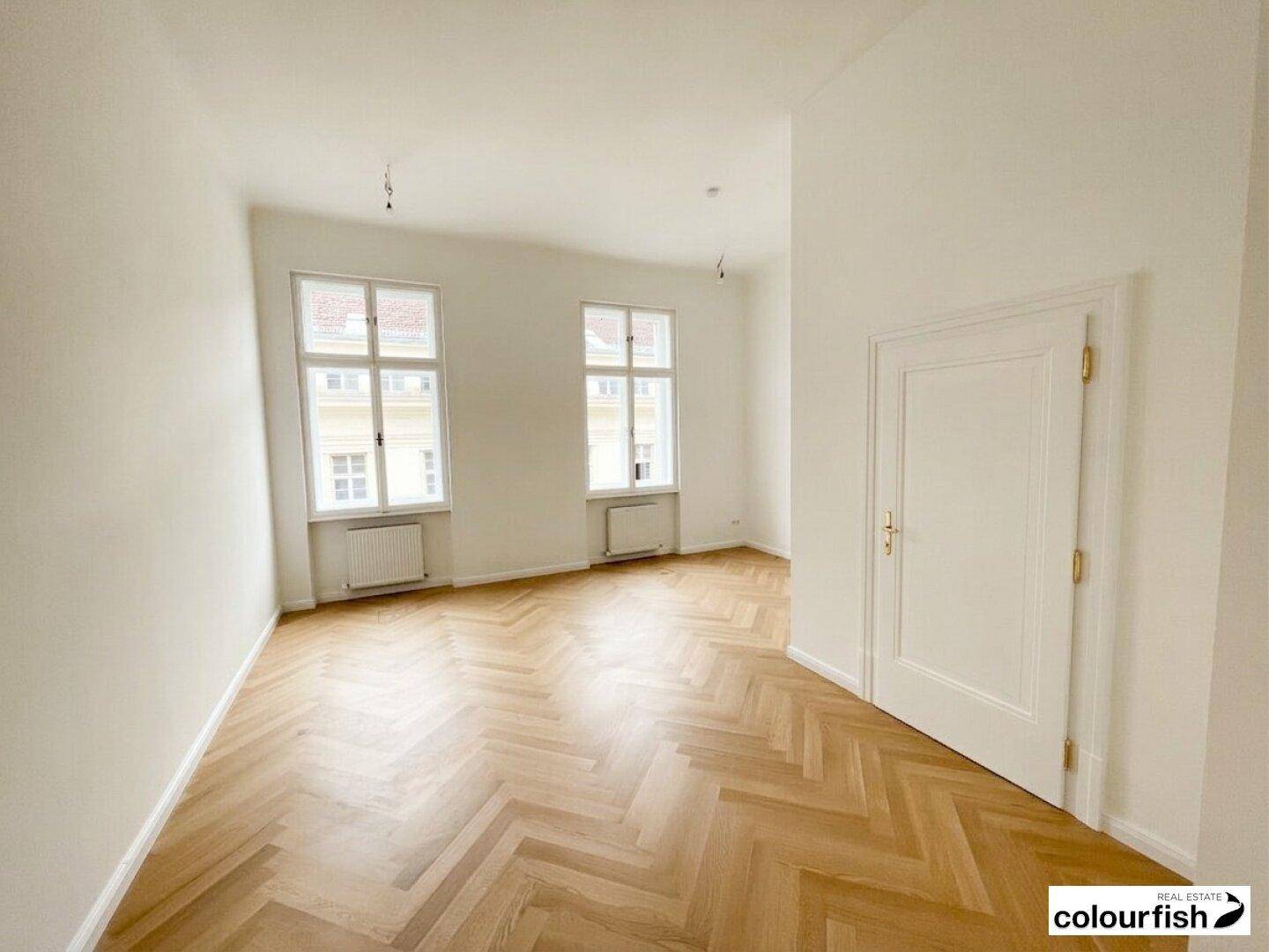 Heller, leerer Raum mit Fischgrätparkett, weißen Wänden und zwei großen Fenstern, vielseitig nutzbar.
