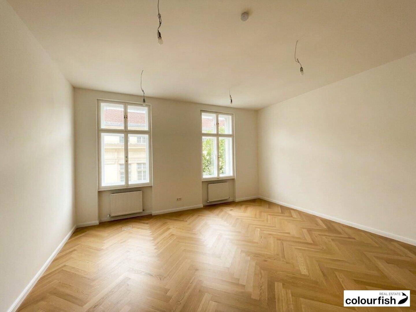 Geräumiger, heller Raum mit Fischgrätparkett, weißen Wänden und zwei großen Fenstern, vielseitig einsetzbar.