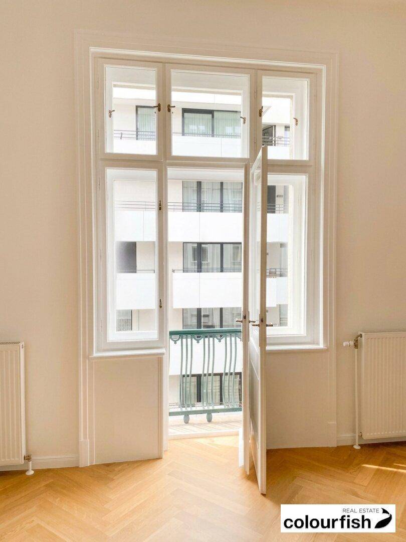 Heller Raum mit Fischgrätparkett und einem großen Fenster, das zu einem französischen Balkon führt.