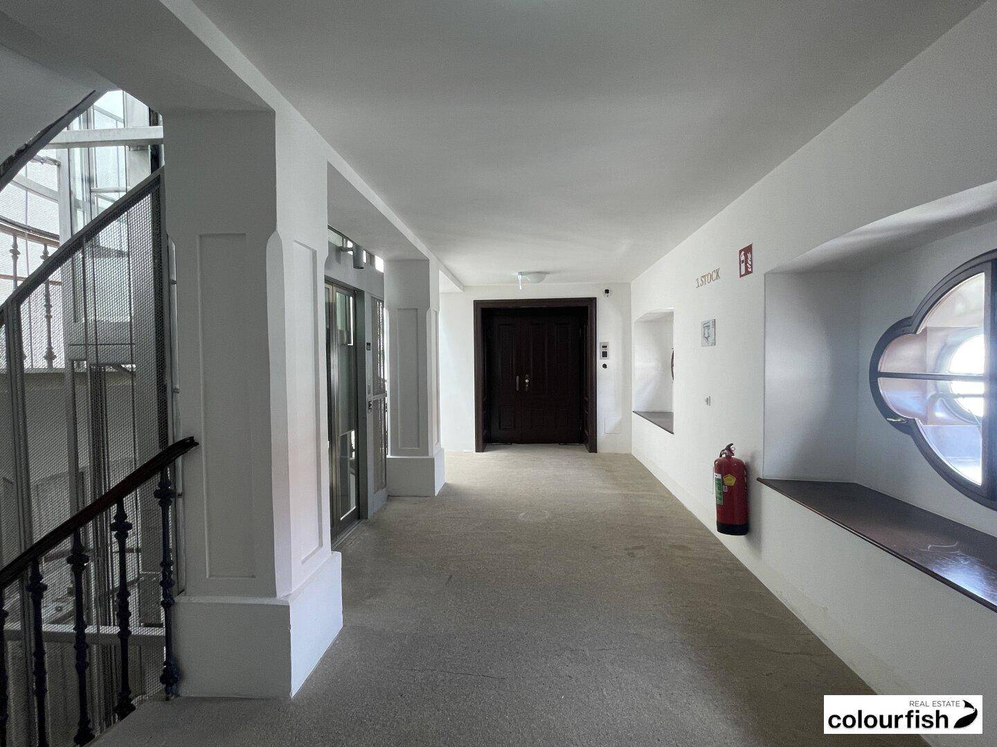 Heller Flur mit modernem Glasaufzug, einer Treppe und einem Feuerlöscher an der Wand.
