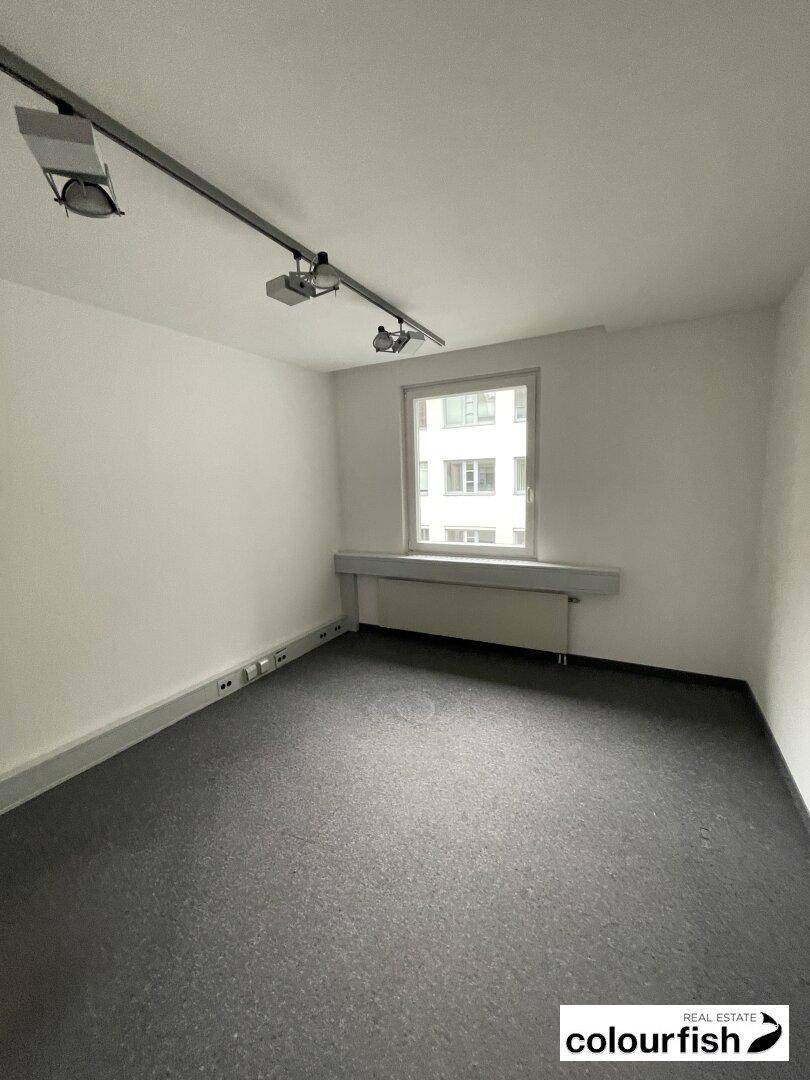 Kleinerer Büroraum mit Teppichboden, einem Fenster und moderner Schienenbeleuchtung.