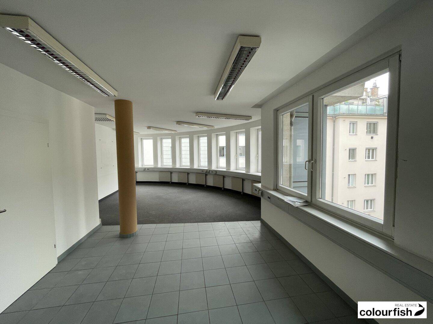 Großer Büroraum mit gemischtem Bodenbelag, einer Säule und einer geschwungenen Fensterfront.