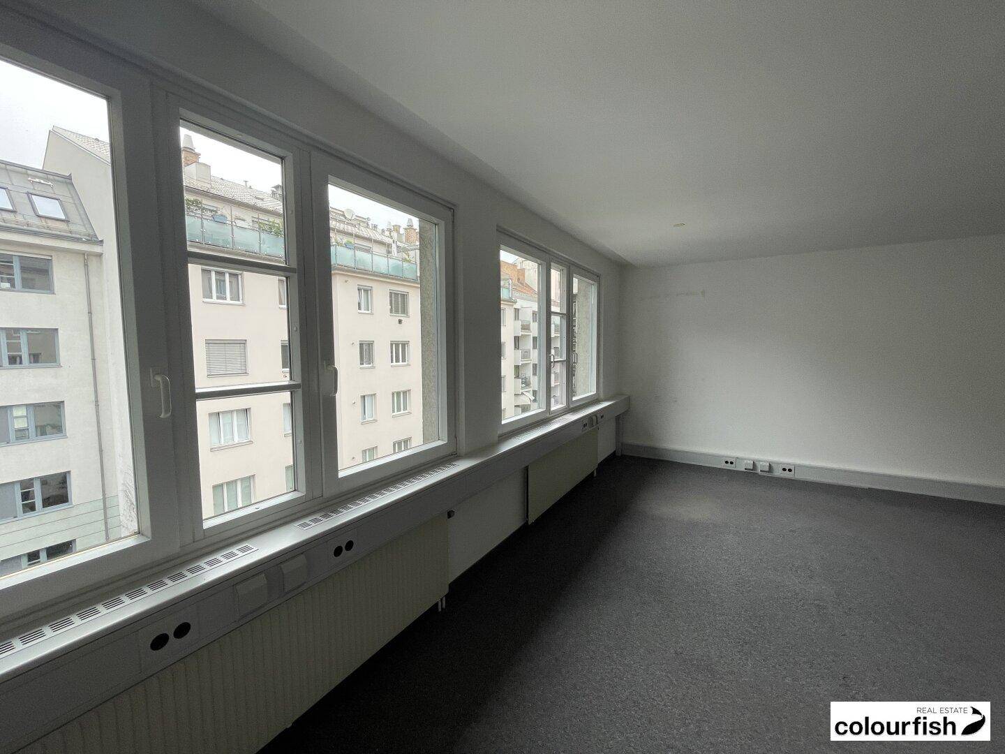Heller und geräumiger Büroraum mit dunklem Teppichboden und einer langen Fensterreihe.