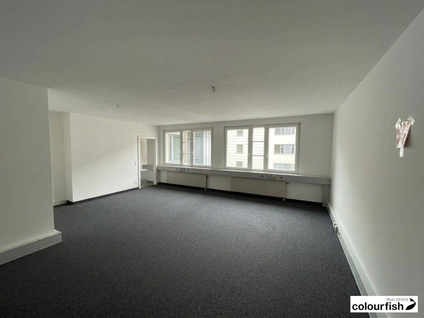Heller Büroraum mit Teppichboden und mehreren Fenstern, die den Raum gut belichten.