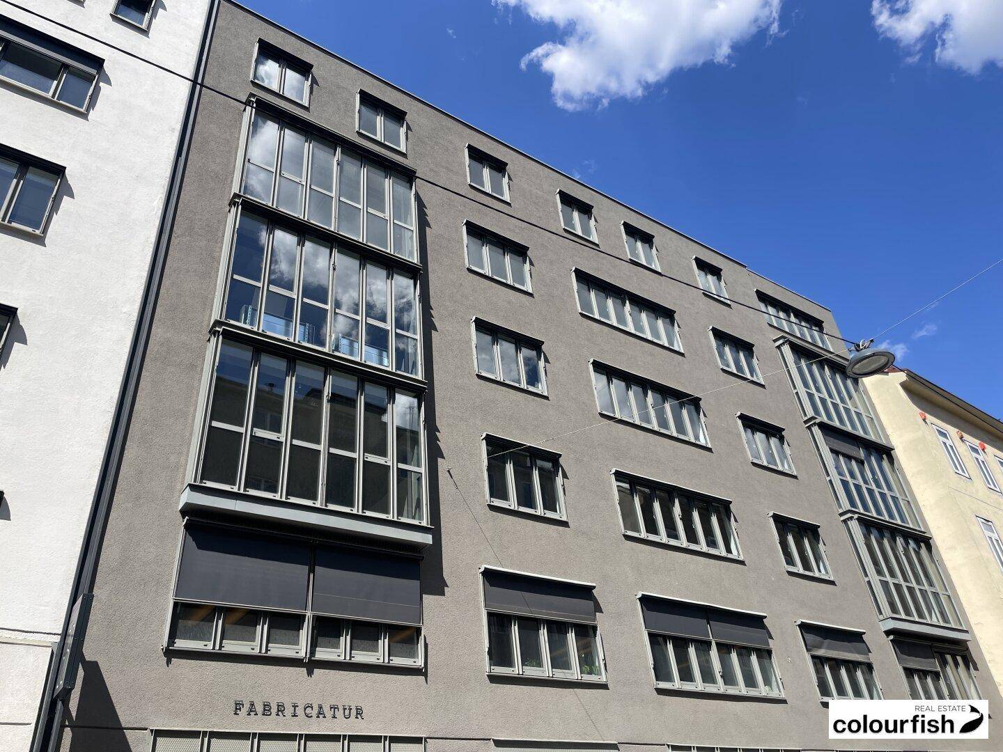 Modernes Gebäude mit vielen Fenstern und einer grauen Fassade unter blauem Himmel.