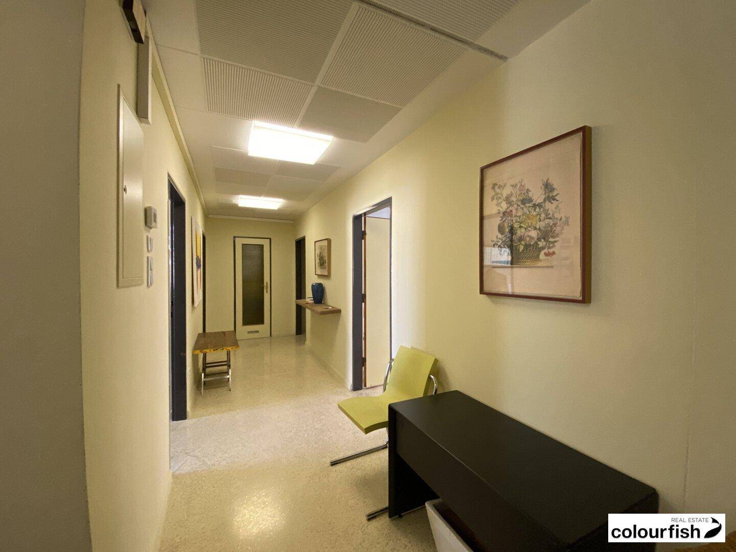 Langer Flur mit hellen Wänden, Deckenbeleuchtung und Bildern an der Wand.