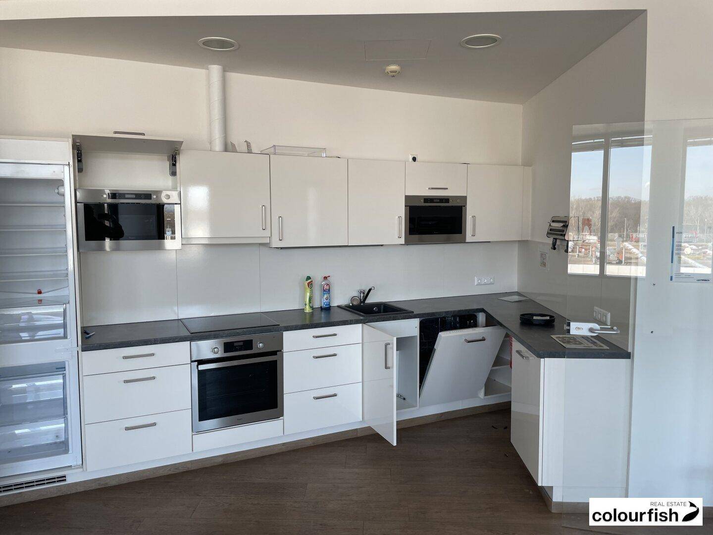 Moderne Einbauküche mit weißen Hochglanzschränken, dunklen Arbeitsplatten und integrierten Geräten, inklusive Fenster.