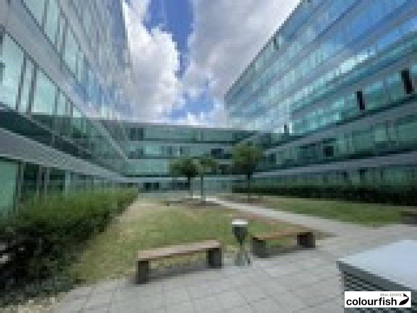 Moderne Bürogebäude mit Glasfassaden und einem begrünten Innenhof mit Sitzbänken unter blauem Himmel.