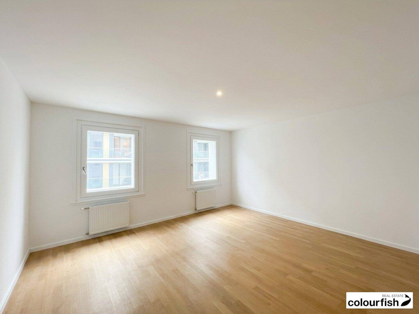 Heller, leerer Raum mit zwei Fenstern, weiß gestrichenen Wänden und einem warmen Holzfußboden.