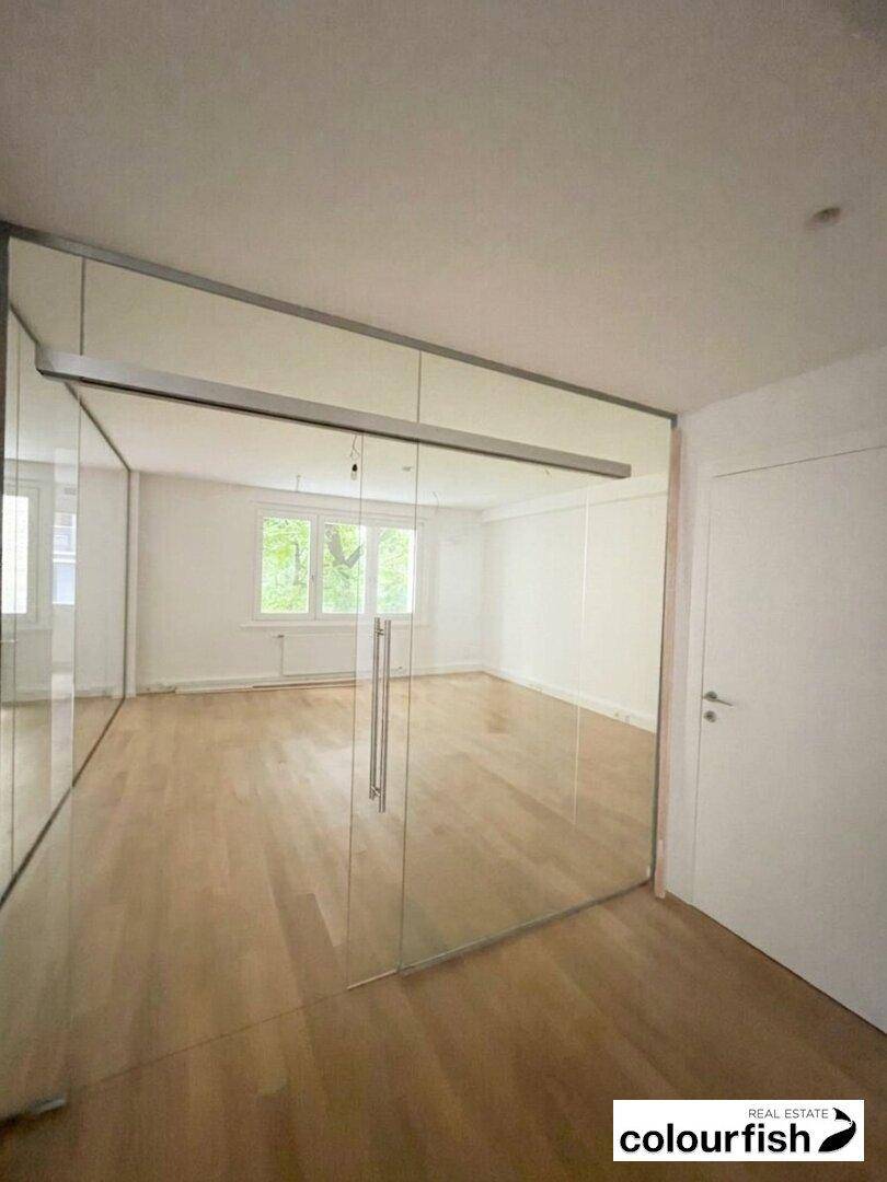 Geräumiger Büroraum mit großen Glaswänden, hellem Holzfußboden und Blick ins Grüne durch die Fenster.